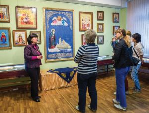 A group of women stand on a wooden floor in a gallery looking at colorful icon paintings. 