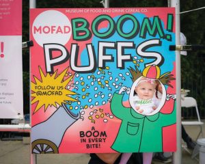View of a picture opportunity at the Museum of Food and Drink with MOFAD Boom Puffs board with a hole in the right side for a person to put their face through. 