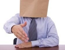 A man with a paper bag over his head extends his hand to greet to the viewer, As if he is entering an interview.