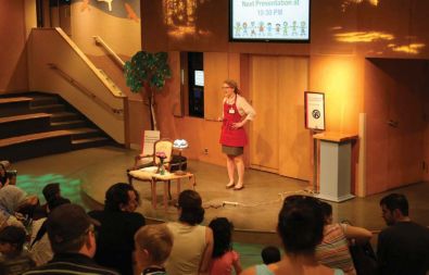 A woman in a red apron stands on a stage in front of a set of doors lecturing an audience of adults and children. 