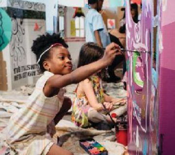 Three children sit on the floor in front of a wall painting. 