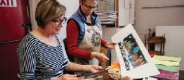 Two woman stand looking at paintings on a table. 