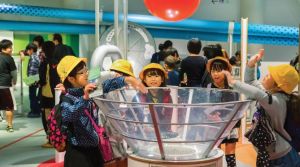 A group of children stand around an air conduit wearing hard hats. 