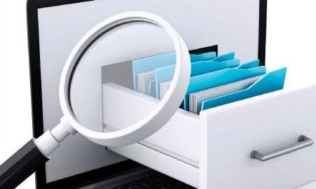 A laptop computer sits open on a desk with a card catalog file drawer coming out of the screen and a magnifying glass looking at the cards.