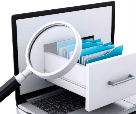 A laptop computer sits open on a desk with a card catalog file drawer coming out of the screen and a magnifying glass looking at the cards.