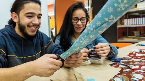 A young hispanic man and a young hispanic woman sit at a table with various wrapping paper smiling and making various crafts.