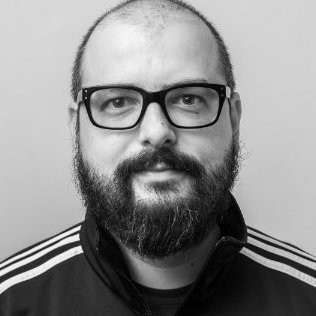 Headshot of a man with a close shaved head, thick dark colored beard and mustache wearing dark rimmed classes.