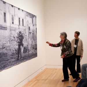 Two women examine a large format photograph on the wall. One of the women is reaching toward the image.