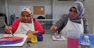 Two women sit facing the camera wearing head scarves and white aprons apparently painting artwork that is sitting on a grey table. 