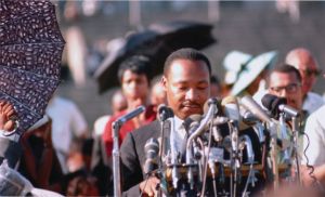 Image of Dr. Martin Luther King, Jr. standing behind a podium filled with microphones and many people milling about behind him. 