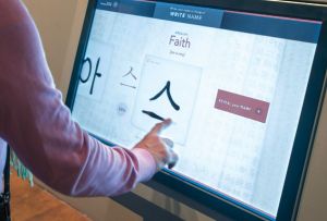 View of someone's arm wearing a long-sleeve pink shirt touching an interactive screen that says Faith above an Asian character beneath it. 