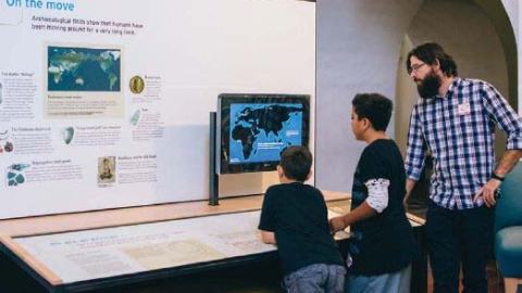 A man and two young boys look at a map on a screen in a museum exhibition.