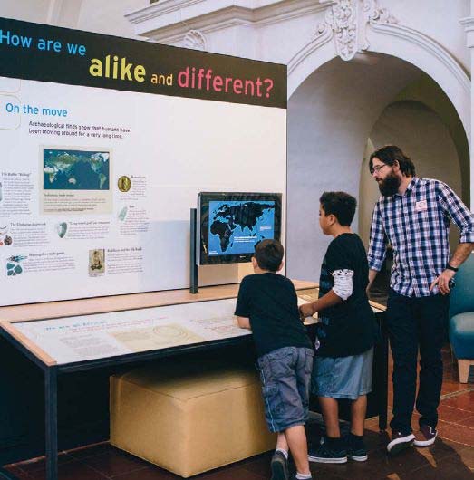 A man and two young boys look at a map on a screen in a museum exhibition.
