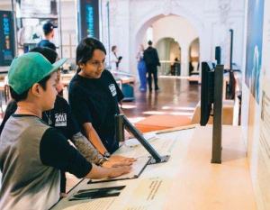 Three young children stand with their hands on an interactive in a museum exhibition.