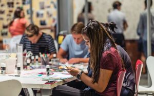 Several people sit at a table working on paper projects with various art supplies in front of them. 