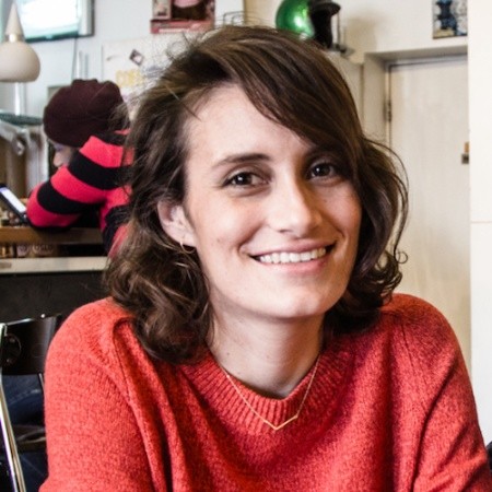 Image of a woman sitting down smiling broadly at the camera wearing a red sweater with someone seated in the distance behind her. She has shoulder length brown hair.