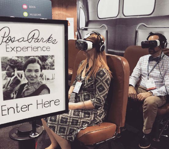 A woman sits in front of a man in a bus like exhibition area wearing VR masks and headphones.
