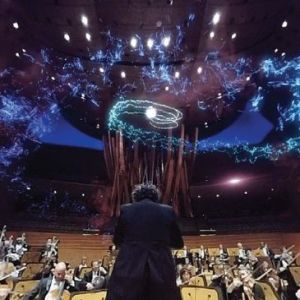 Image of a male conductor standing in front of an orchestra on a raised platform with a blue colored light display shimmering in the air above their heads.