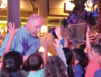 Phil Manning shown speaking in front of a group of young children with their hands raised. 