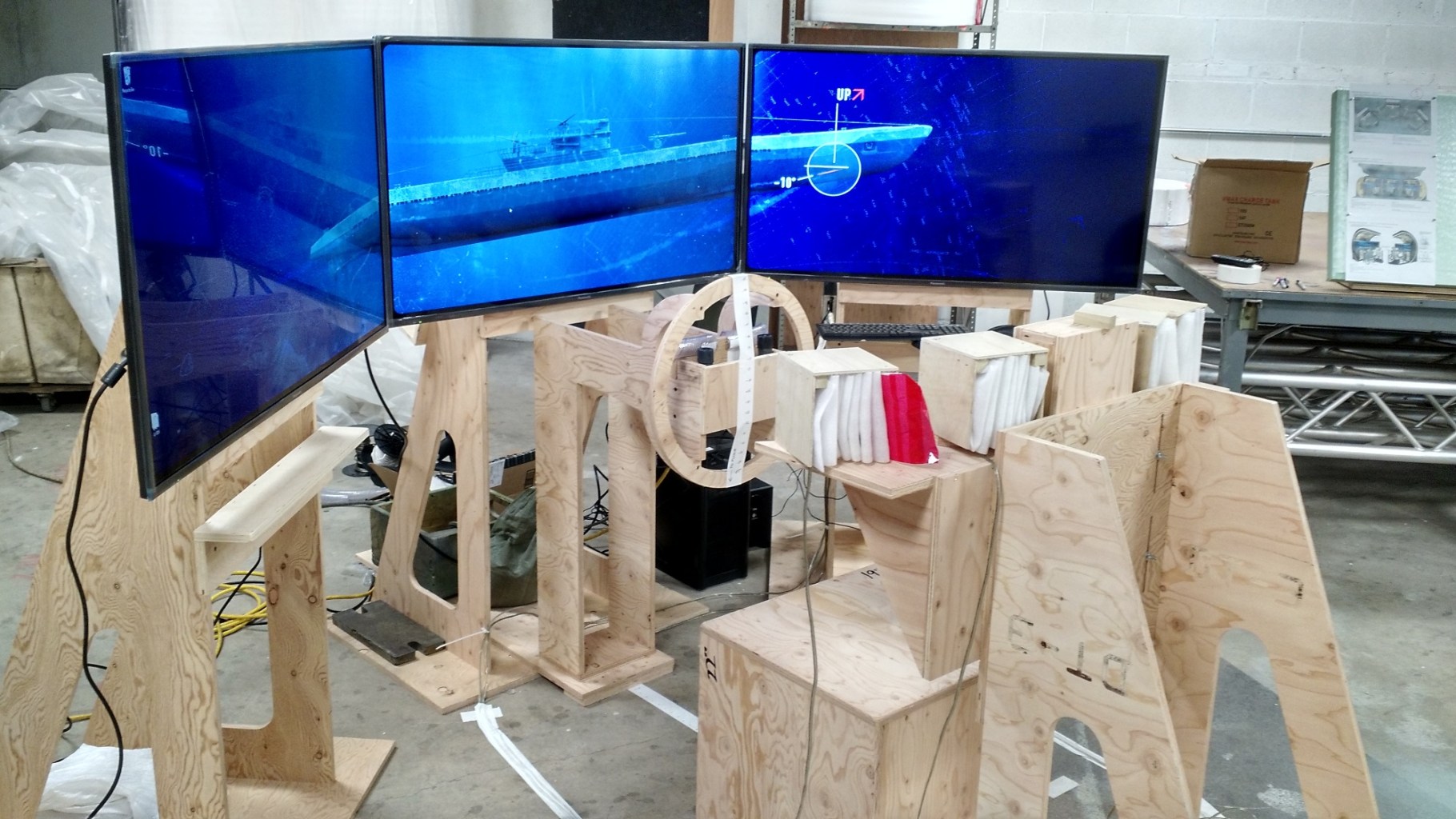 An image of three LCD screens in a semi circle standing on saw horses in a workshop with the image of a submarine displayed on the screens. 