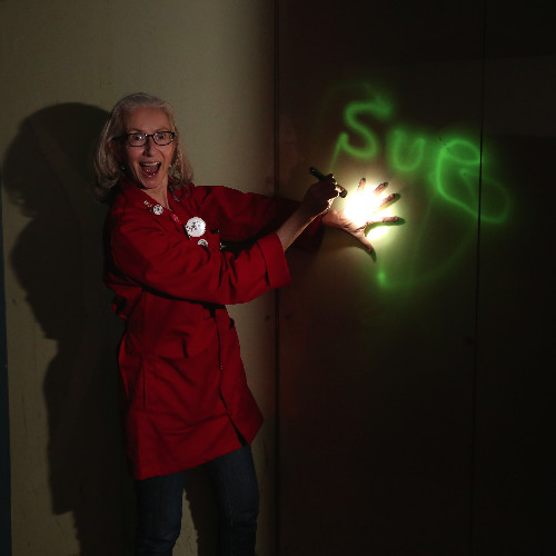 A woman stands next to a light shining on a wall with green letters spelling out Sue. She looks extremely excited with her mouth open, wearing a red lab coat.