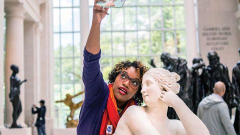 A black woman stands behind a Grecian statue taking a selfie.