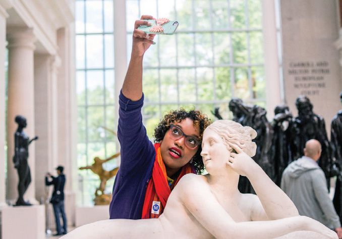 A black woman stands behind a Grecian statue taking a selfie.