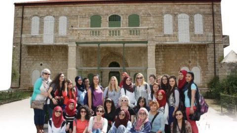 A large group of people stand and sit in two rows in front of a Spanish inspired building.