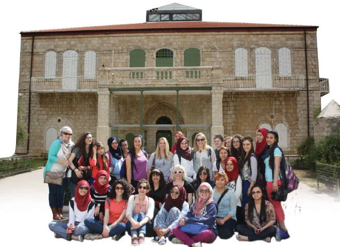 A large group of people stand and sit in two rows in front of a Spanish inspired building.