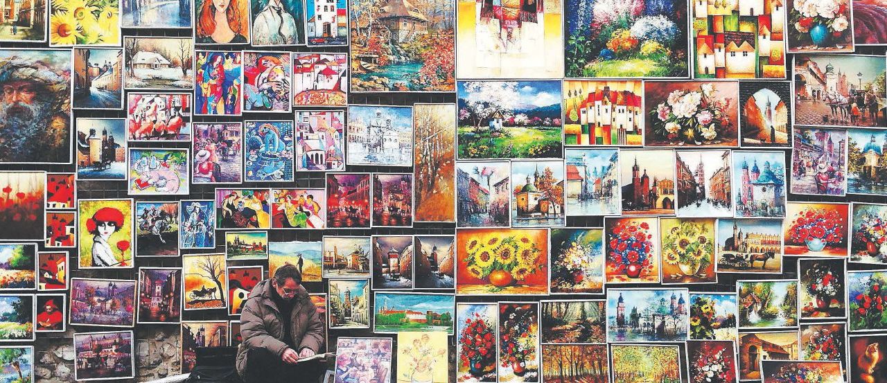 A man crouches in front of a large wall with a variety of oil paintings hanging on it.