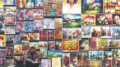 A man crouches in front of a large wall with a variety of oil paintings hanging on it.