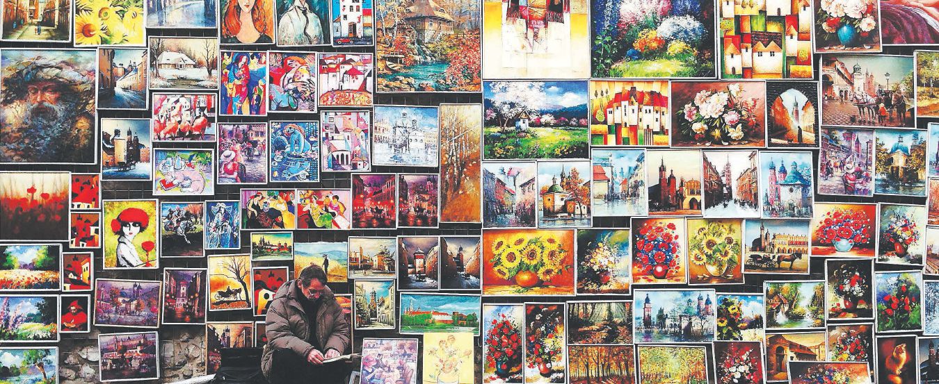 A man crouches in front of a large wall with a variety of oil paintings hanging on it.