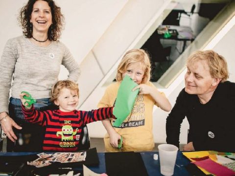 A young family looks towards the camera. Mom is smiling broadly with shoulder length curly hair,. Two young boys interact with paper materials in front of them, each holds a pair of scissors one has a piece of paper in front of his face. The father looks on from a seated position wearing a black long sleeve shirt, he has medium length blond hair.