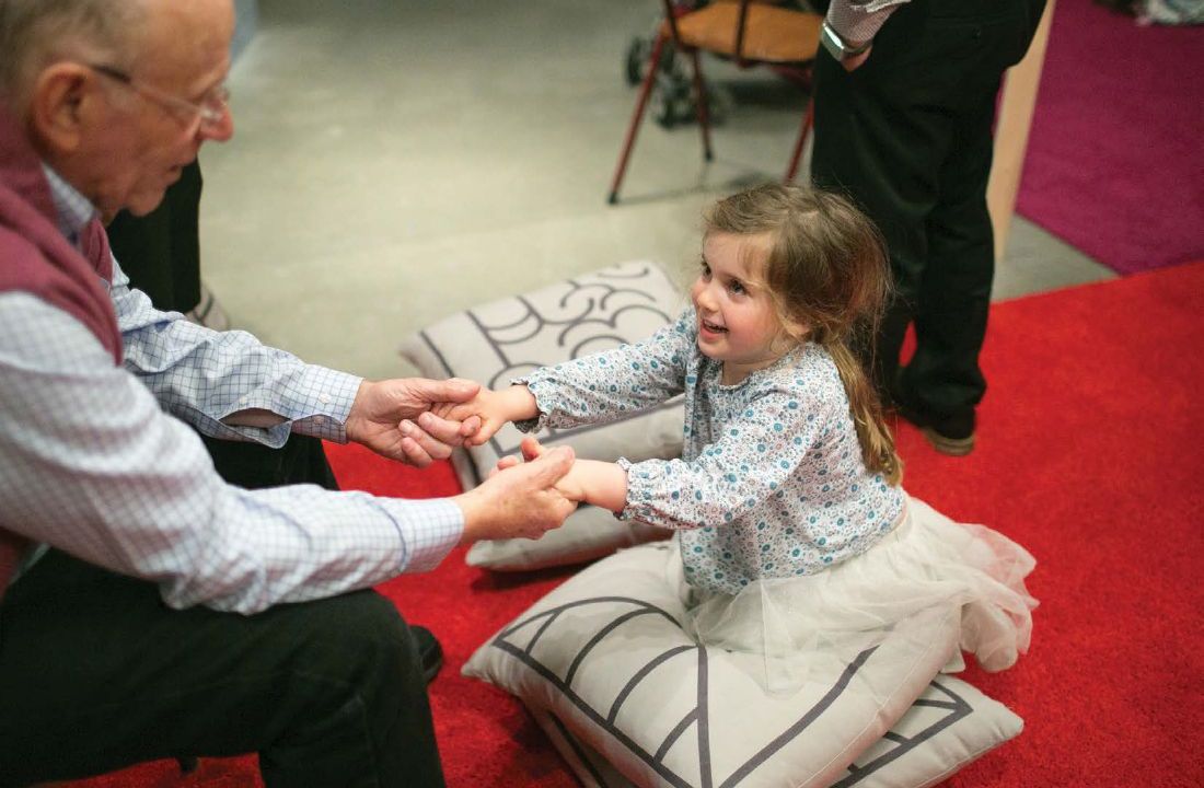 A little girl in a patterned shirt and tutu skirt kneels on a set of pillows facing an older gentleman who is clasping her hands and smiling.