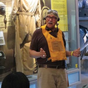 A grey haired man wearing a deflated flotation device stands in front of a museum case and appears to be giving a tour. 