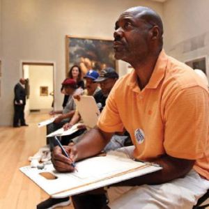A man sitting in an art gallery stares intently at a painting (off-screen) with an art clipboard and pen. 