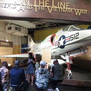 A female tour guide stands in front of a fighter jet with a group of uniformed people in front of her.