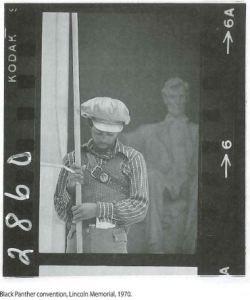 Image of a black and white photo negative of an African American man standing outside the Lincoln memorial holding a long pole. 