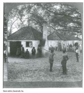 Historical black and white photograph of several African American people standing and leaning against a few small white brick structures. 