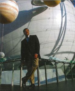 Neil deGrasse Tyson leaning on a metal railing with a few planets behind. 