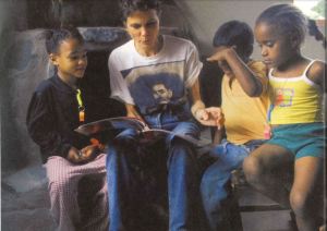 Several young children sit next to a woman who is reading out load with a book in her lap. 