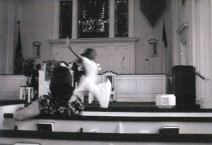 A woman is dancing and leaping in the front of a church congregation. 