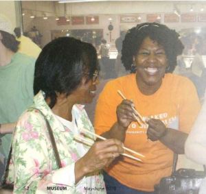 Two women hold chopsticks while smiling broadly at the camera in a crowded market. 