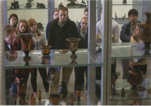 A group of people stand looking into a display case. The camera shot is taken through the display case from the other side. 