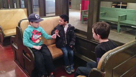 Three boys sit in a train car exhibited in a gallery.