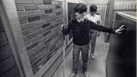 Two young men walk down a hallway with various wall treatments (for example brick, concrete) wearing blindfolds and carrying walking canes that a person with blindness might use.