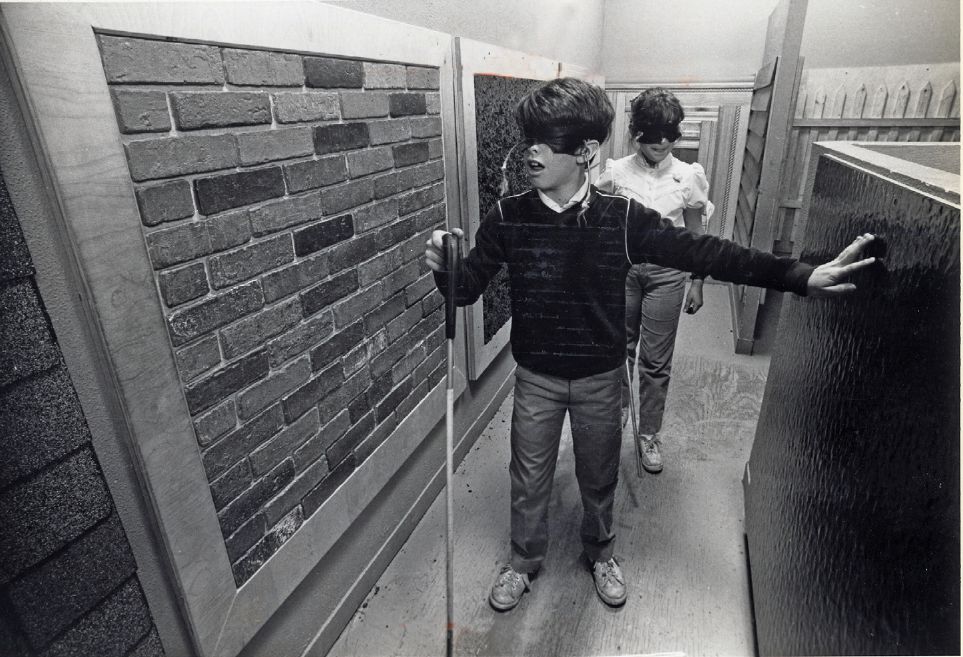 Two young men walk down a hallway with various wall treatments (for example brick, concrete) wearing blindfolds and carrying walking canes that a person with blindness might use.