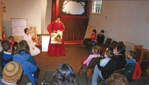 A costumed interpreter stands in front of a group of young people showing them an art project. 