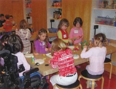 Children sitting around a table with a young girl in wheelchair at one end working on an art project. 