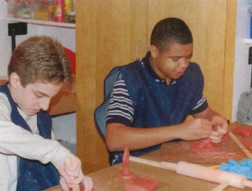 Two young men sit working side-by-side at a table working on an art project with clay. 